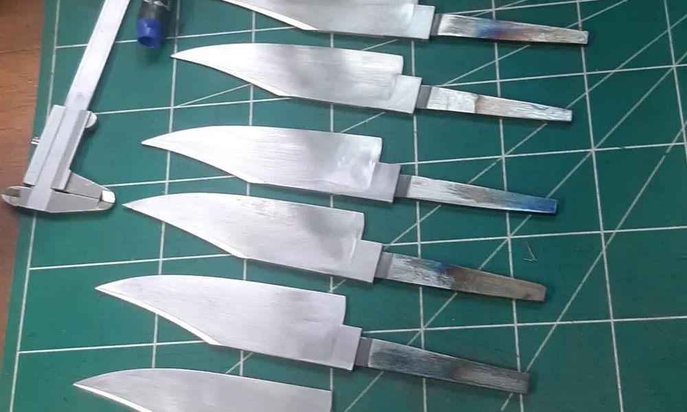Bastidores de produção: lâminas artesanais em bruto após forjamento, ainda sem cabo, mostrando as cores de têmpera do aço