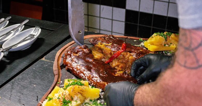 Faca artesanal cravada em tábua de madeira com carne assada e mandioca em restaurante — Runefall Cutelaria