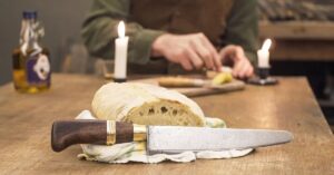 Faca artesanal com cabo em madeira escura e guarda em latão apoiada em pão rústico sobre mesa de madeira com velas — Runefall Cutelaria