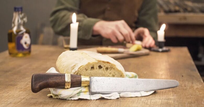 Faca artesanal com cabo em madeira escura e guarda em latão apoiada em pão rústico sobre mesa de madeira com velas — Runefall Cutelaria