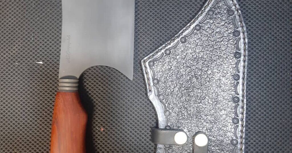 Faca artesanal com cabo curvado ergonômico e bainha de couro trabalhada, exemplo de acabamento premium Runefall