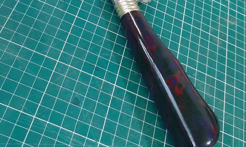 Cabo de faca artesanal em Micarta com padrão marmorizado vermelho e azul, exemplo de material moderno usado como alternativa à madeira