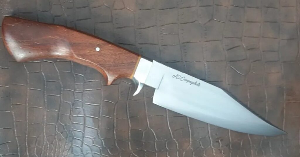 Faca de caça artesanal com lâmina em aço 5160, guarda e pino em inox, separador em resina e cabo em jatobá do campo, sobre fundo em couro texturizado — Runefall Cutelaria